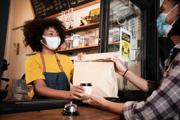 African American female barista with face mask works in the cafe with social distance, takeaway coffee for a customer, new normal service of small business coffee shop in COVID19 quarantine lifestyle. - Photo, image