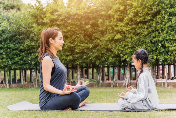 Asian young mother practicing doing yoga exercises with her daughter outdoors in meditate pose together on green grass in nature a field garden park, family sport and exercises for healthy lifestyle - Photo, image