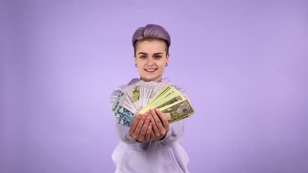 Joyful young adult woman generation Z with short purple hair holding bunch of different currencies, tossing them up, rejoicing, boasting money profit. Indoor studio shot isolated on purple background - Footage, Video