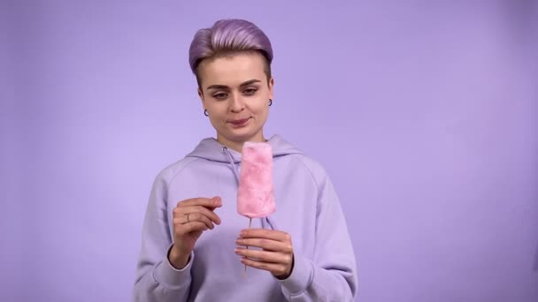 Vrolijke jonge volwassen vrouw met suikerspin op stok, kijken naar zoet goed in afwachting, likken lippen, krijgen stuk van snoep floss, eten met plezier geïsoleerd op paarse achtergrond binnen - Video