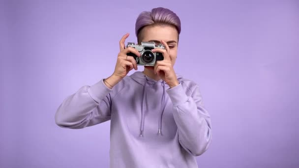 Cheerful young adult woman with trendy look taking pictures with old retro film camera, turning advance lever photographing with joy isolated on purple background indoors. Charming girl wearing hoodie - Footage, Video