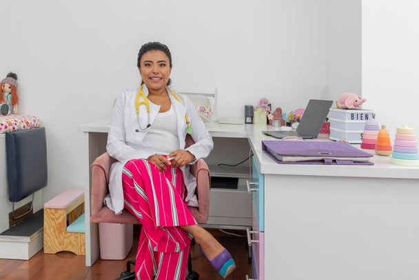 Latina naispuolinen lastenlääkäri istuu pöydällään stetoskoopilla. - Valokuva, kuva