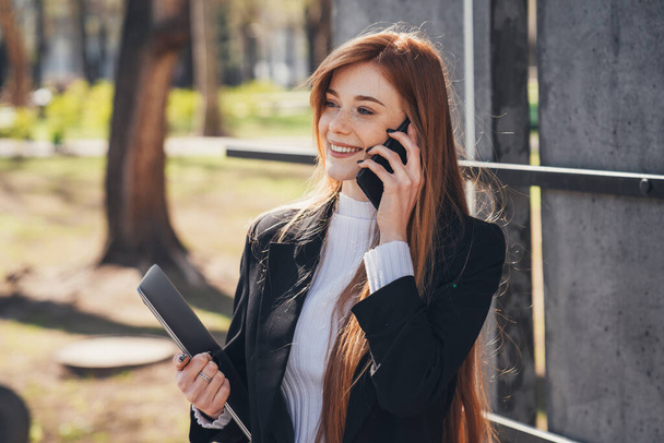 Modern red-haired businesswoman talking on phone sitting on bench in spring park outdoors, holding laptop. People urban lifestyle concep. Female using phone. - Foto, afbeelding