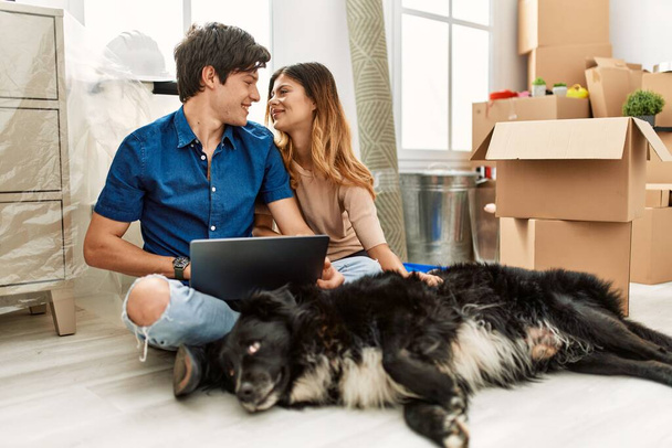Genç beyaz çift yeni evlerinde köpekleriyle yerde otururken dizüstü bilgisayar kullanıyorlar.. - Fotoğraf, Görsel