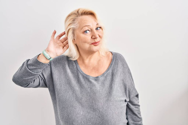 Middle age caucasian woman standing over white background smiling with hand over ear listening an hearing to rumor or gossip. deafness concept.  - Valokuva, kuva