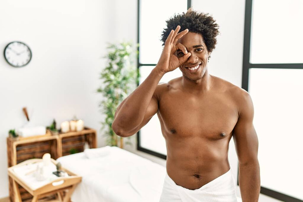 Jovem homem americano africano sem camisa usando toalha de pé no centro de beleza fazendo gesto ok com a mão sorrindo, olho olhando através dos dedos com o rosto feliz.  - Foto, Imagem