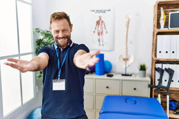 Middle age physiotherapist man working at pain recovery clinic looking at the camera smiling with open arms for hug. cheerful expression embracing happiness.  - Zdjęcie, obraz
