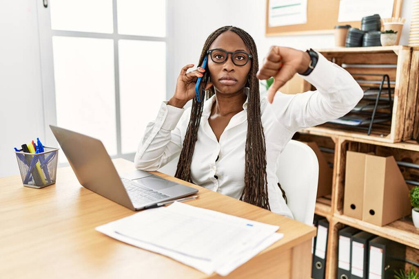 Czarna kobieta z warkoczami pracująca w biurze, mówiąca przez telefon, wyglądająca na nieszczęśliwą i wściekłą, wykazująca odrzucenie i negatyw z opuszczonymi kciukami. złe wyrażenie.  - Zdjęcie, obraz