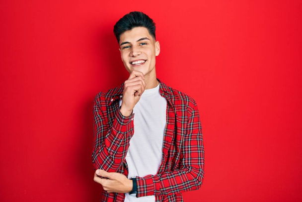 Jonge Spaanse man met casual kleding die zelfverzekerd naar de camera kijkt met een glimlach met gekruiste armen en hand geheven op de kin. positief denken.  - Foto, afbeelding