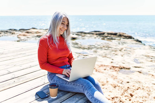 Medioevo donna dai capelli grigi utilizzando computer portatile seduto sulla panchina in spiaggia. - Foto, immagini