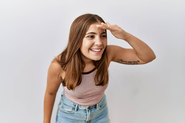 Young brunette woman standing over isolated background very happy and smiling looking far away with hand over head. searching concept.  - Foto, imagen