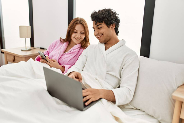 Молода пара посміхається щасливий, використовуючи ноутбук і смартфон у спальні
. - Фото, зображення