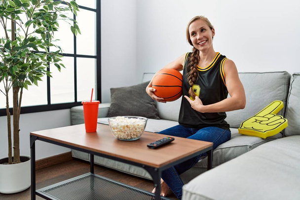 Νεαρή ξανθιά γυναίκα κρατώντας μπάλα του μπάσκετ ζητωκραυγάζοντας παιχνίδι τηλεόραση χαμογελώντας χαρούμενος δείχνοντας με το χέρι και το δάχτυλο  - Φωτογραφία, εικόνα