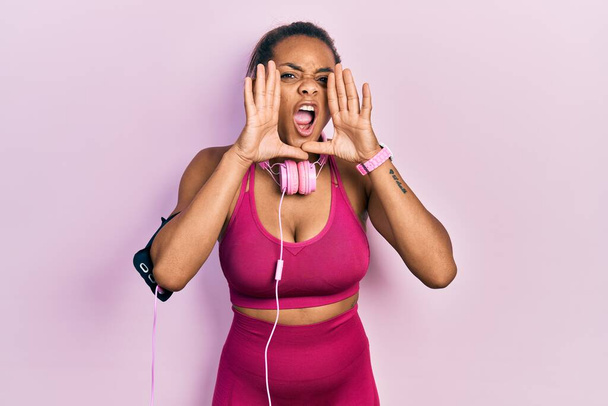 Giovane ragazza afroamericana indossa abiti da palestra e utilizzando cuffie urlando arrabbiato ad alta voce con le mani sulla bocca  - Foto, immagini