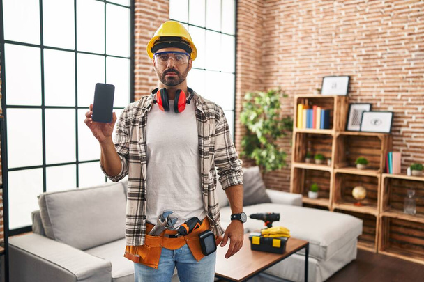 Молодий іспаномовний чоловік з бородою працює вдома, тримаючи смартфон мислення ставлення і тверезий вираз, дивлячись на себе впевнено
  - Фото, зображення