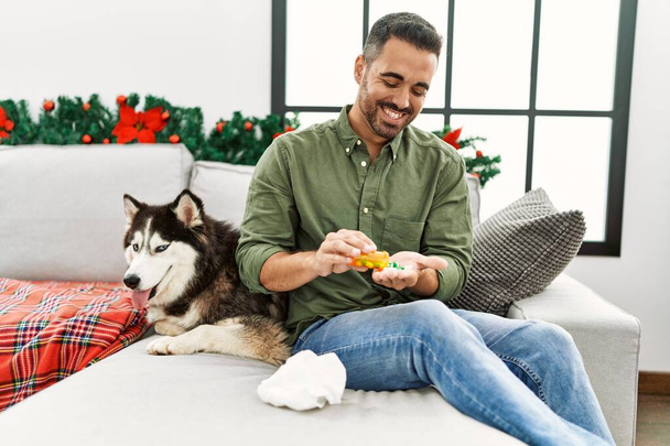 młody Hiszpan biorąc pigułki siedzi na kanapie z psem na Boże Narodzenie wystrój w domu - Zdjęcie, obraz