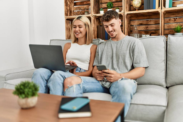 Νεαρό καυκάσιο ζευγάρι χαμογελά χαρούμενος χρησιμοποιώντας φορητό υπολογιστή και smartphone κάθεται στον καναπέ στο σπίτι. - Φωτογραφία, εικόνα