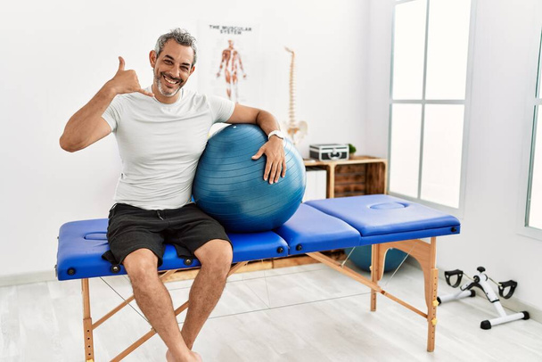 Middle age hispanic man at pain recovery clinic holding pilates ball smiling doing phone gesture with hand and fingers like talking on the telephone. communicating concepts.  - Photo, Image
