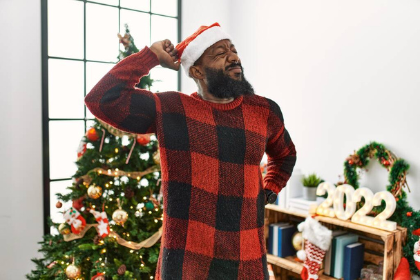 Αφροαμερικάνος που φοράει το καπέλο του Άη Βασίλη στέκεται δίπλα στο χριστουγεννιάτικο δέντρο τεντωμένο πίσω, κουρασμένος και χαλαρός, νυσταγμένος και χασμουριέται νωρίς το πρωί  - Φωτογραφία, εικόνα