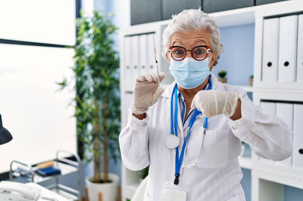 Ηλικιωμένη γυναίκα με γκρίζα μαλλιά φορώντας ιατρική στολή και ιατρική μάσκα κρατώντας σύριγγα δείχνοντας το δάχτυλο σε έναν εαυτό χαμογελαστός χαρούμενος και περήφανος  - Φωτογραφία, εικόνα