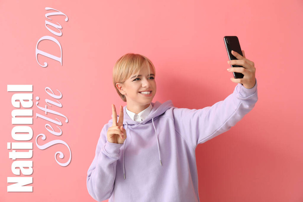 Mooie jonge vrouw in stijlvolle hoodie het nemen van selfie op roze achtergrond - Foto, afbeelding