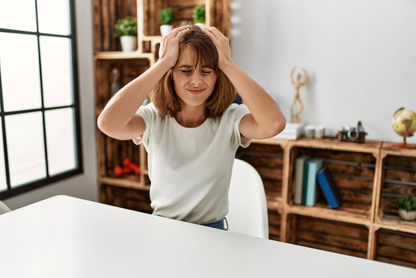 Junge kaukasische Mädchen in lässiger Kleidung sitzen auf dem Tisch zu Hause unter Kopfschmerzen verzweifelt und gestresst, weil Schmerzen und Migräne. Hände auf den Kopf.  - Foto, Bild