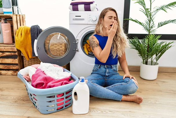 Young beautiful woman doing laundry sitting by wicker basket bored yawning tired covering mouth with hand. restless and sleepiness.  - Photo, Image