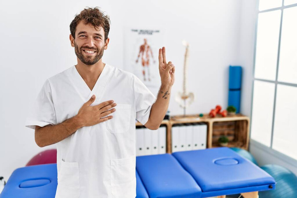 Молодой красивый физиотерапевт, работающий в клинике болеутоляющих, улыбаясь, ругаясь рукой на груди и пальцах вверх, давая клятву верности  - Фото, изображение