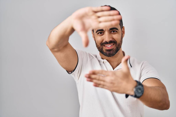 Молодий іспаномовний чоловік з бородою носить повсякденний одяг на білому тлі, посміхаючись роблячи рамку руками і пальцями з щасливим обличчям. креативність і концепція фотографії
.  - Фото, зображення