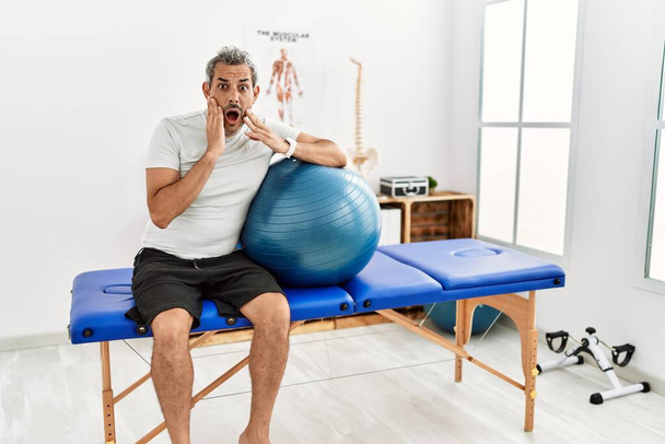 Middle age hispanic man at pain recovery clinic holding pilates ball afraid and shocked, surprise and amazed expression with hands on face  - Photo, Image