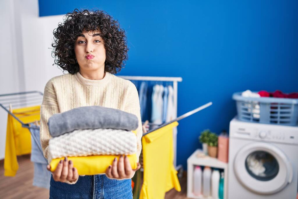 Young brunette woman with curly hair holding clean laundry puffing cheeks with funny face. mouth inflated with air, catching air.  - Photo, Image