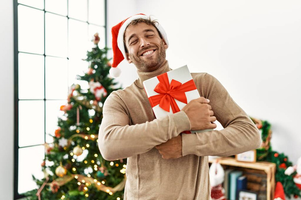 Joven hombre hispano sonriendo confiado sosteniendo regalo de Navidad en casa - Foto, imagen
