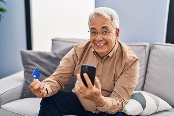 Hombre mayor usando teléfono inteligente y tarjeta de crédito sentado en el sofá en casa - Foto, Imagen