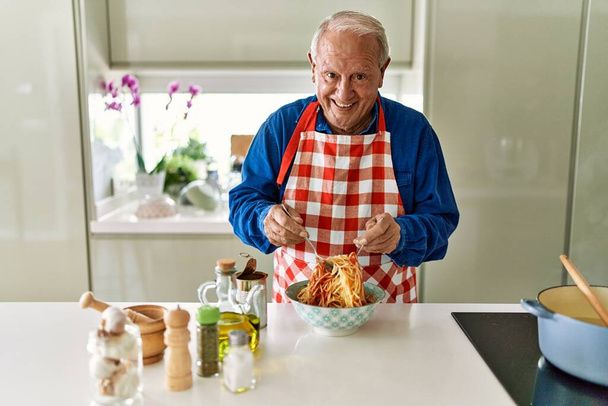 Senior άνθρωπος χαμογελά αυτοπεποίθηση ανάμειξη σάλτσα ντομάτας με μακαρόνια στην κουζίνα - Φωτογραφία, εικόνα