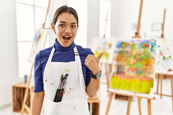 Giovane donna bruna in studio d'arte sorpreso indicando con il dito della mano di lato, bocca aperta espressione stupita.  - Foto, immagini