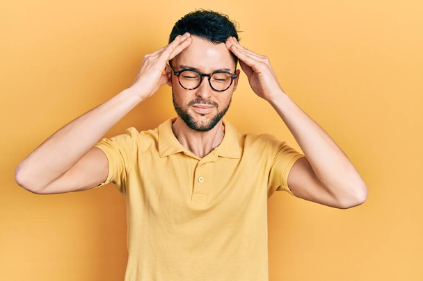 Fiatal spanyol férfi alkalmi ruhát és szemüveget visel, kezével a fején, hogy fejfájást okozzon a stressz. migrénben szenved.  - Fotó, kép