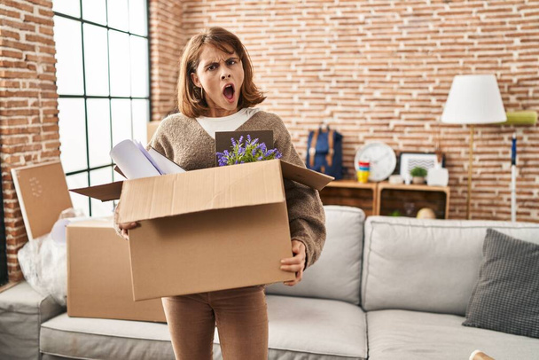 Νεαρή όμορφη γυναίκα κρατώντας κουτί κινείται σε ένα νέο σπίτι στο πρόσωπο σοκ, αναζητούν δύσπιστοι και σαρκαστικός, έκπληκτος με ανοιχτό στόμα  - Φωτογραφία, εικόνα