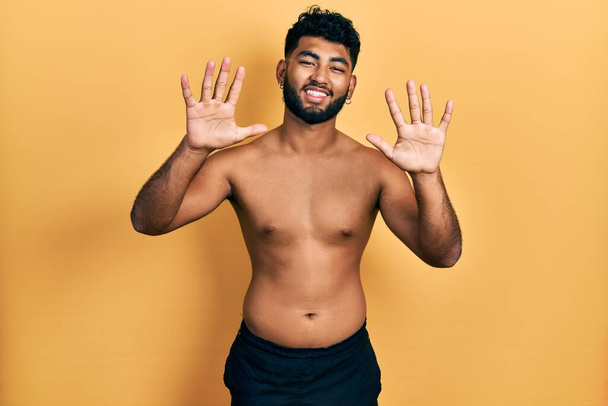 Arabischer Mann mit Bart trägt Badehosen und zeigt mit Finger Nummer zehn nach oben, während er selbstbewusst und glücklich lächelt.  - Foto, Bild