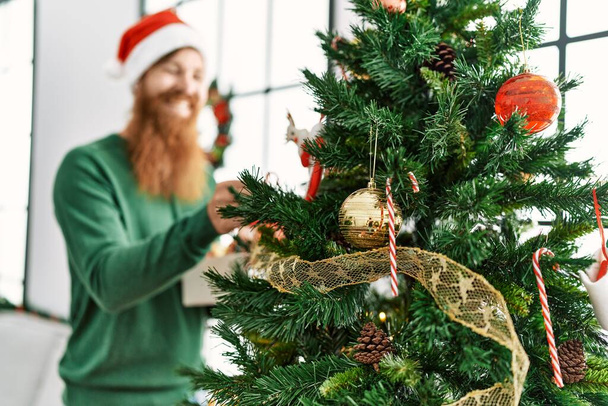 Junger Rotschopf lächelt selbstbewusst und schmückt den Weihnachtsbaum zu Hause - Foto, Bild