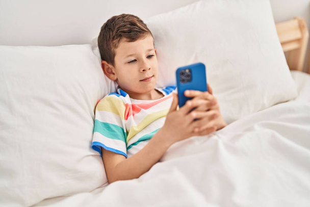 Enfant blond utilisant un smartphone couché sur le lit dans la chambre - Photo, image