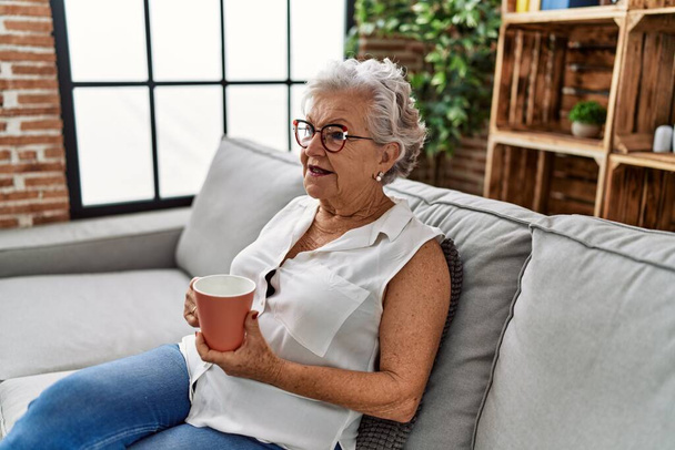 Yaşlı gri saçlı kadın evdeki kanepede kahve içiyor. - Fotoğraf, Görsel