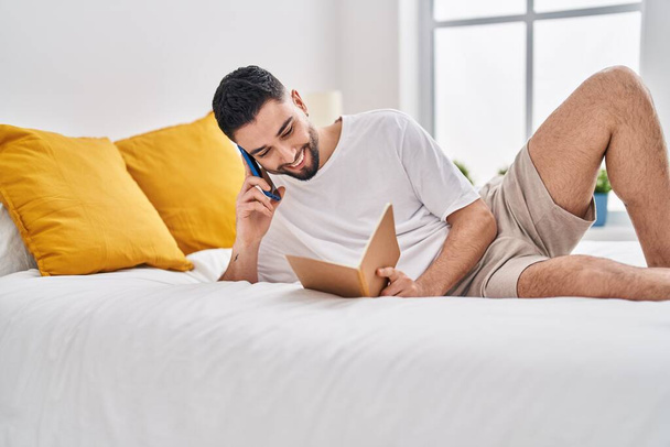 Hombre árabe joven hablando en el libro de lectura de teléfonos inteligentes en el dormitorio - Foto, imagen