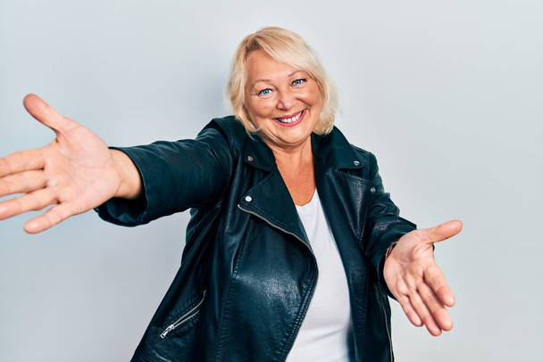 Middle age blonde woman wearing leather jacket looking at the camera smiling with open arms for hug. cheerful expression embracing happiness.  - 写真・画像