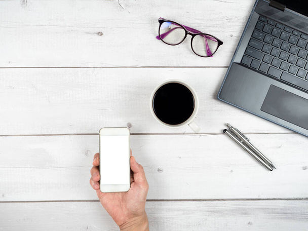 Ноутбук очки кофе чашки и две серебряные ручки офис рабочее пространство вид сверху и рука держать смартфон белый экран копирования пространство - Фото, изображение