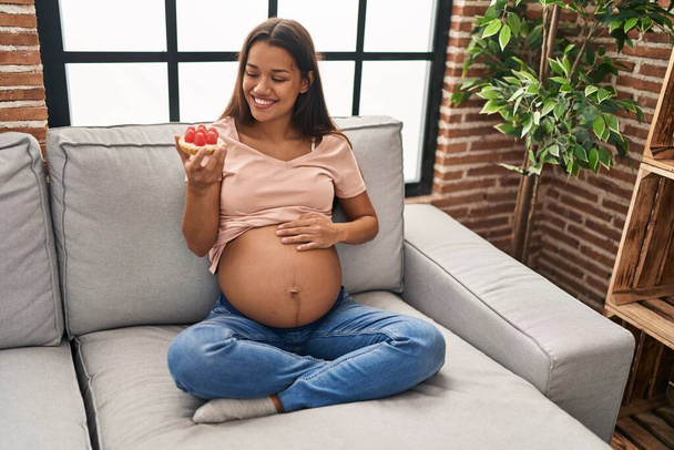 Jovem mulher latina grávida comer bolo sentado no sofá em casa - Foto, Imagem
