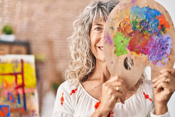 Femme d'âge moyen tenant palette peintre couvrant le visage souriant avec un sourire heureux et cool sur le visage. montrant les dents.  - Photo, image