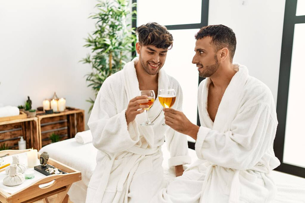 Two hispanic men couple toasting with champagne sitting on massage table at beauty center - Zdjęcie, obraz