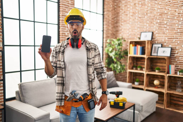 Junger hispanischer Mann mit Bart bei Renovierungsarbeiten zu Hause, Smartphone in der Hand, erschrocken und erstaunt mit offenem Mund für Überraschung, ungläubiges Gesicht  - Foto, Bild