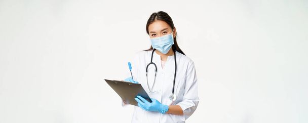 Улыбающаяся азиатка-врач в маске и перчатках, записывающая диагноз пациента, рецепт, ручка и буфер обмена, белый фон. - Фото, изображение