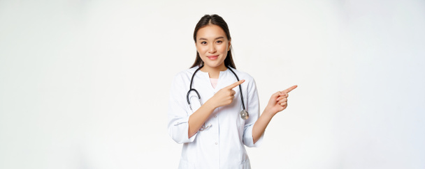 Assistenza sanitaria, concetto di persone mediche. Sorridente asiatica medico femminile, indicando le dita a destra, mostrando pubblicità, ospedale informazioni, in piedi su sfondo bianco. - Foto, immagini